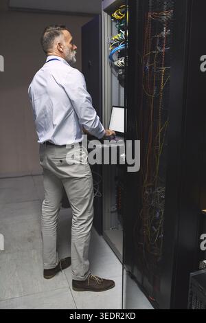 Typing, cybersecurity and man on laptop in server room for diagnostics, inspection and network. Database, engineer and mature person on computer for Stock Photo