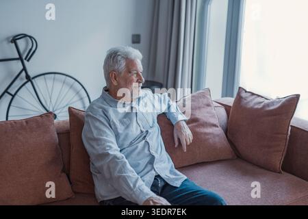 An older man sits in his living room looking through the window with a serene expression, reflecting on his day and appreciating a quiet and restful moment at home Stock Photo
