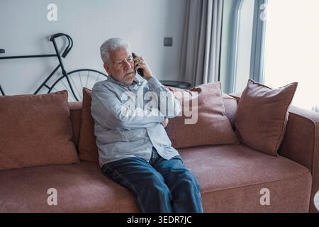 Worried concerned senior elder man talking on cellphone to doctor, feeling unwell, calling for emergency, ambulance, sitting on sofa at home, contact helpline, using mobile phone Stock Photo