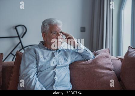 Elderly man sits alone on a sofa looking outside with a sad expression. He seems stressed and reflective while dealing with personal concerns at home. Stock Photo