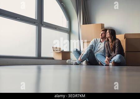 A happy married couple in their 30s rests on a sofa on moving day, hugging and looking into the distance while dreaming about a bright future in their first home, with cardboard boxes nearby symbolizing relocation and a new beginning Stock Photo