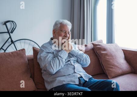 An elderly man sits on a sofa at home holding his cheek in pain, suffering from a toothache and showing discomfort while dealing with dental problems in a quiet living room setting Stock Photo