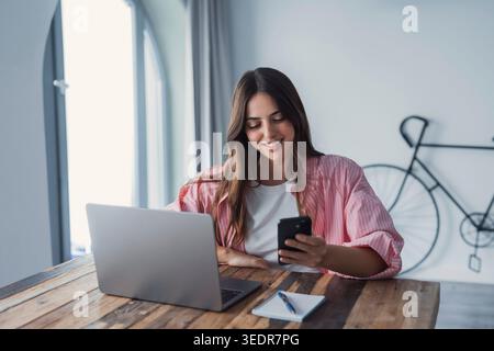 A focused young female entrepreneur works remotely, communicating via online messaging and mobile applications while handling tasks on her laptop in a home office Stock Photo