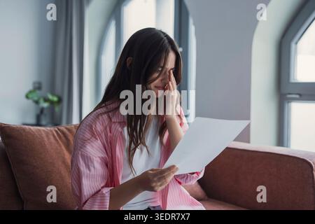 A focused young woman sits on a sofa at home, reading a university acceptance letter and studying official documents, carefully analyzing course details and academic requirements Stock Photo