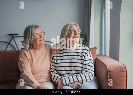Senior women sitting on sofa at home looking away through window, peaceful and thoughtful elderly females enjoying quiet moment together, friendship, reflection, active aging and calm lifestyle concept Stock Photo