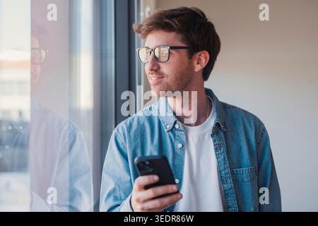 Dreamy young Hispanic businessman in office, holding smartphone and using app, smiling and looking out the window, showing thoughtful and positive expression Stock Photo