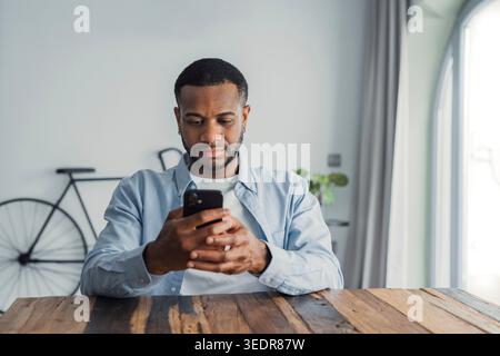 Confused African guy holding smart phone feels concerned thinking over received message. Mobile phone everyday usage, unpleasant news, waiting for important call, low signal device problems concept Stock Photo