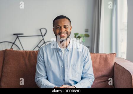 Head shot close up smiling handsome young african ethnicity biracial cheerful man looking at camera, having fun enjoying video call conversation with friends, resting on comfortable sofa at home. Stock Photo