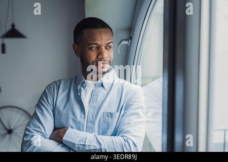 Thoughtful millennial African American man looking at window away in deep thoughts, thinking over problems, bad news, difficult tasks, feeling bored, sad. Portrait of pensive guy, homeowner Stock Photo