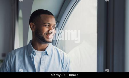 Smiling dreamy African American man thinking about future or new opportunities, positive pensive businessman at window looking to aside, planning, pondering strategy, business vision concept Stock Photo