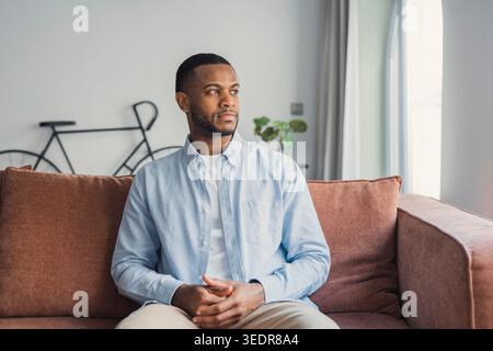 Thoughtful bored millennial Black mixed race guy lost in thoughts, sitting on coach, looking away, feeling frustration, boredom, apathy, laziness, thinking over bad news, problems, difficult tasks Stock Photo