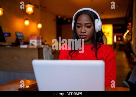 Young black woman concentrating on laptop, wearing headphones in a warm lit cafe Stock Photo