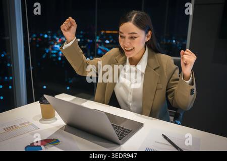 success businesswoman with arm raised while working with laptop computer in business high-rise office with city lights background at night Stock Photo