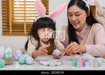 mother and preschool child girl wearing bunny ears painting and decorating egg  together at their home on easter day. happy family holiday activity co Stock Photo