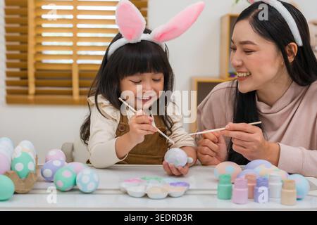 mother and preschool child girl wearing bunny ears painting and decorating egg  together at their home on easter day. happy family holiday activity co Stock Photo