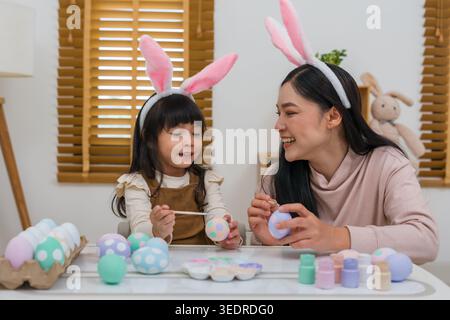mother and preschool child girl wearing bunny ears painting and decorating egg  together at their home on easter day. happy family holiday activity co Stock Photo