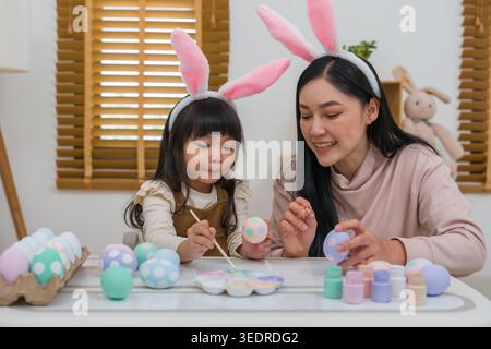 mother and preschool child girl wearing bunny ears painting and decorating egg  together at their home on easter day. happy family holiday activity co Stock Photo