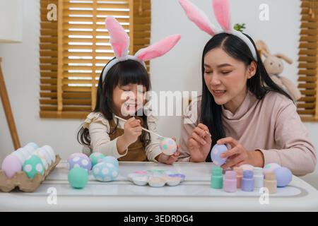 mother and preschool child girl wearing bunny ears painting and decorating egg  together at their home on easter day. happy family holiday activity co Stock Photo