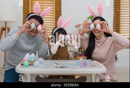 father, mother and daughter wearing bunny ears close their eyes with egg and laughing at their home on easter day. happy family holiday activity conce Stock Photo