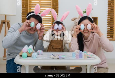 father, mother and daughter wearing bunny ears close their eyes with egg and laughing at their home on easter day. happy family holiday activity conce Stock Photo