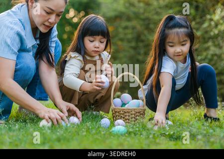 mother and her two daughters playing and hunting for Easter eggs into a basket on green grass in the garden Stock Photo