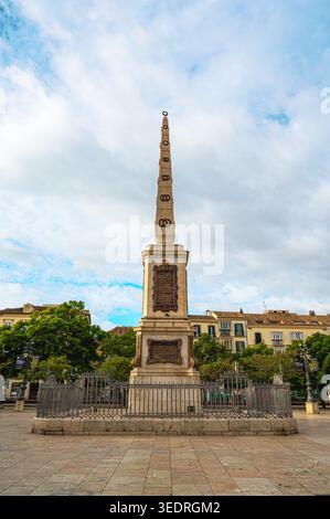 Tall stone obelisk monument on a stone pedestal surrounded by a wrought iron fence in a paved urban square, with historic residential buildings and tr Stock Photo