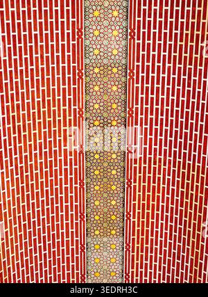 Close-up of a red patterned wall featuring a vertical strip of colorful geometric mosaic tiles. Symmetrical brick-like motifs frame the intricate flor Stock Photo