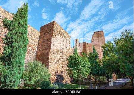 Ancient stone fortress walls rise above a small garden with trees, shrubs, and a paved path under a blue sky with scattered clouds. The warm stone tex Stock Photo