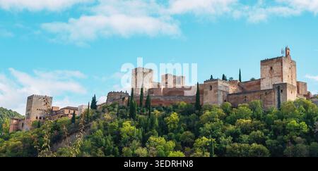 Wide panoramic view of the Alhambra fortress complex on a wooded hill with stone towers and walls under a bright blue sky with scattered clouds in Gra Stock Photo