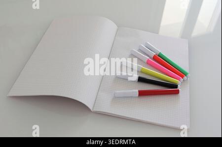 Colorful marker pens are arranged on a blank open squared notebook, symbolizing creativity, education, learning, and new beginnings on a clean white desk Stock Photo