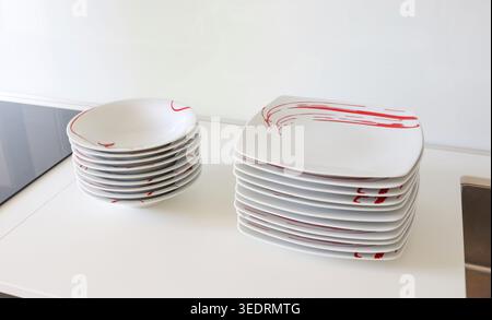 Stacks of clean modern white ceramic dishes with bold red abstract brushstroke patterns, neatly stacked on a kitchen counter ready for serving or table setting preparation Stock Photo
