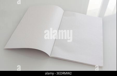 Blank pages of a large grid paper notebook, lying open on a reflective white table surface, offer ample copy space, symbolizing new ideas, learning, and fresh beginnings Stock Photo