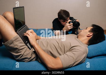 Programmer relaxing on sofa while coding on laptop, another man taking candid photos of him with modern digital camera during casual at-home Stock Photo