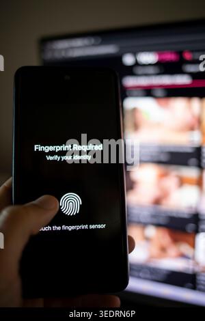 Hand holding a smartphone displaying identity verification by fingerprint, accessing a website requiring age confirmation for adult content shown blurred in the background on a monitor Stock Photo