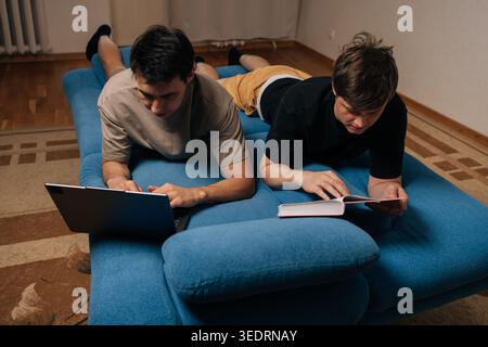 Two young men lying side by side on blue sofa, one actively typing on laptop and other engrossed in reading a book, both demonstrating focused Stock Photo