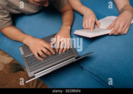 Top view cropped shot of two students studying together on a blue sofa at home, one using a laptop for online learning, the other reading a physical Stock Photo
