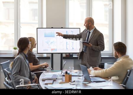 Adult businessman presenting analytics dashboard to colleagues during office meeting, pointing at screen and holding tablet. Ideal for business strategy, teamwork, data reporting Stock Photo