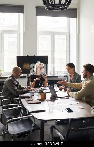 Wide shot of diverse business team discussing strategy during office meeting, collaborating with laptops and documents. Ideal for corporate teamwork, planning, leadership marketing Stock Photo