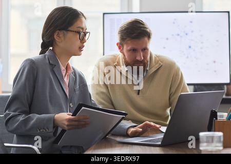 Asian young businesswoman discussing data with adult businessman, reviewing laptop and documents in office. Ideal for teamwork, analytics, strategy content Stock Photo