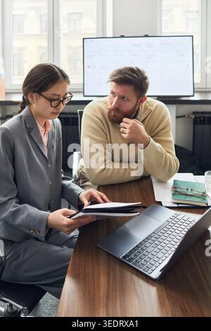Vertical shot of Asian young businesswoman reviewing documents with young businessman during office meeting, supporting teamwork, planning, consulting content Stock Photo