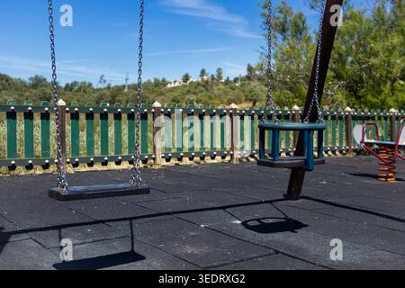 Two empty swings hang from chains in a children's play area, set against a backdrop of a green fence and lush vegetation under a clear blue sky Stock Photo