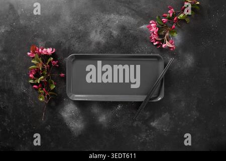 Minimalist scene of empty rectangular plate for asian food with black chopsticks on dark textured surface with spring pink flowers. Asian style. View from above. Copy space. Japanese food concept. Stock Photo