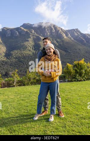 Asian couple standing embracing on lawn at mountain foothills, wearing knit sweater beanie scarf. Romance, outdoors, pair, alpine, ridge, scenery, greenery Stock Photo