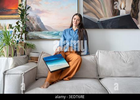 Happy talented female painter posing at camera with artwork, sitting on couch at home loft gallery holding canvas in hand. Positive smiling woman artist poses with painting in front of wall exhibition Stock Photo