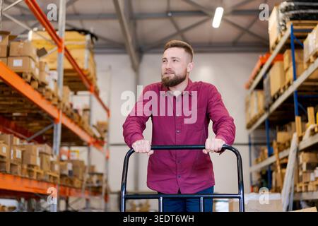 Positive male logistics supervisor in a red shirt standing with a platform trolley. Professional warehouse employee managing distribution and stock delivery among tall storage racks. Stock Photo