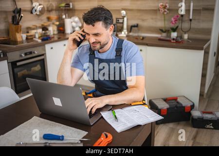 Handyman wearing blue overall using laptop and talking on phone in kitchen Stock Photo