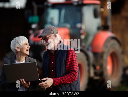 Mature farmers man and woman standing with laptop on farm in front of tractor Stock Photo