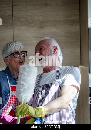 Mature couple having fun while doing chores in kitchen together and singing with duster as microphone Stock Photo