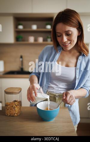 A woman in a kitchen pours milk into a bowl with cereal. She smiles while preparing her breakfast at home in the morning. Stock Photo