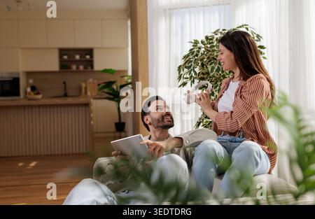 A man sits on a couch holding a tablet while a woman stands nearby with headphones. They are in a sunny living room with plants. Stock Photo